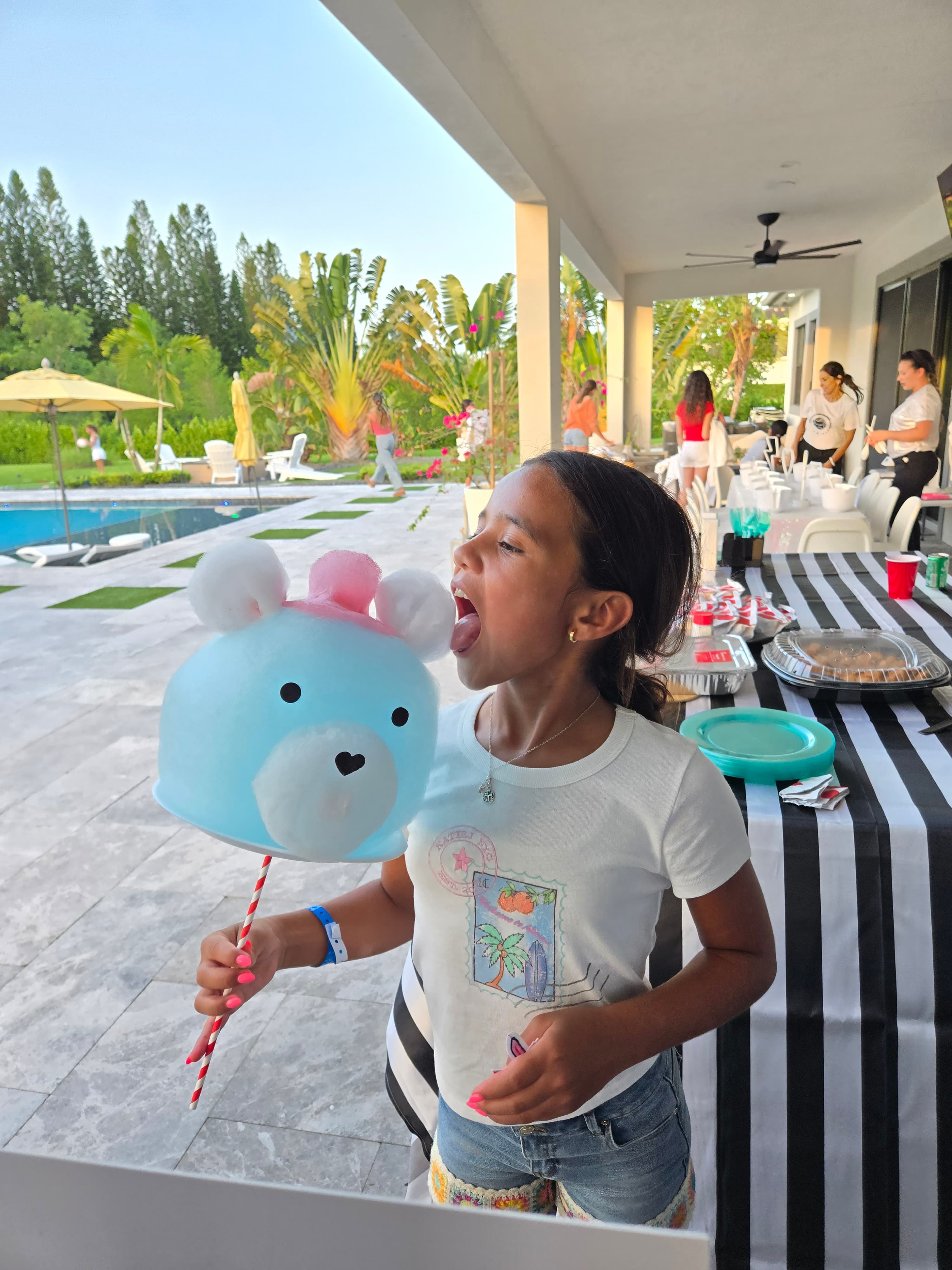 A young girl licks a large, blue bear-shaped cotton candy at an outdoor party.