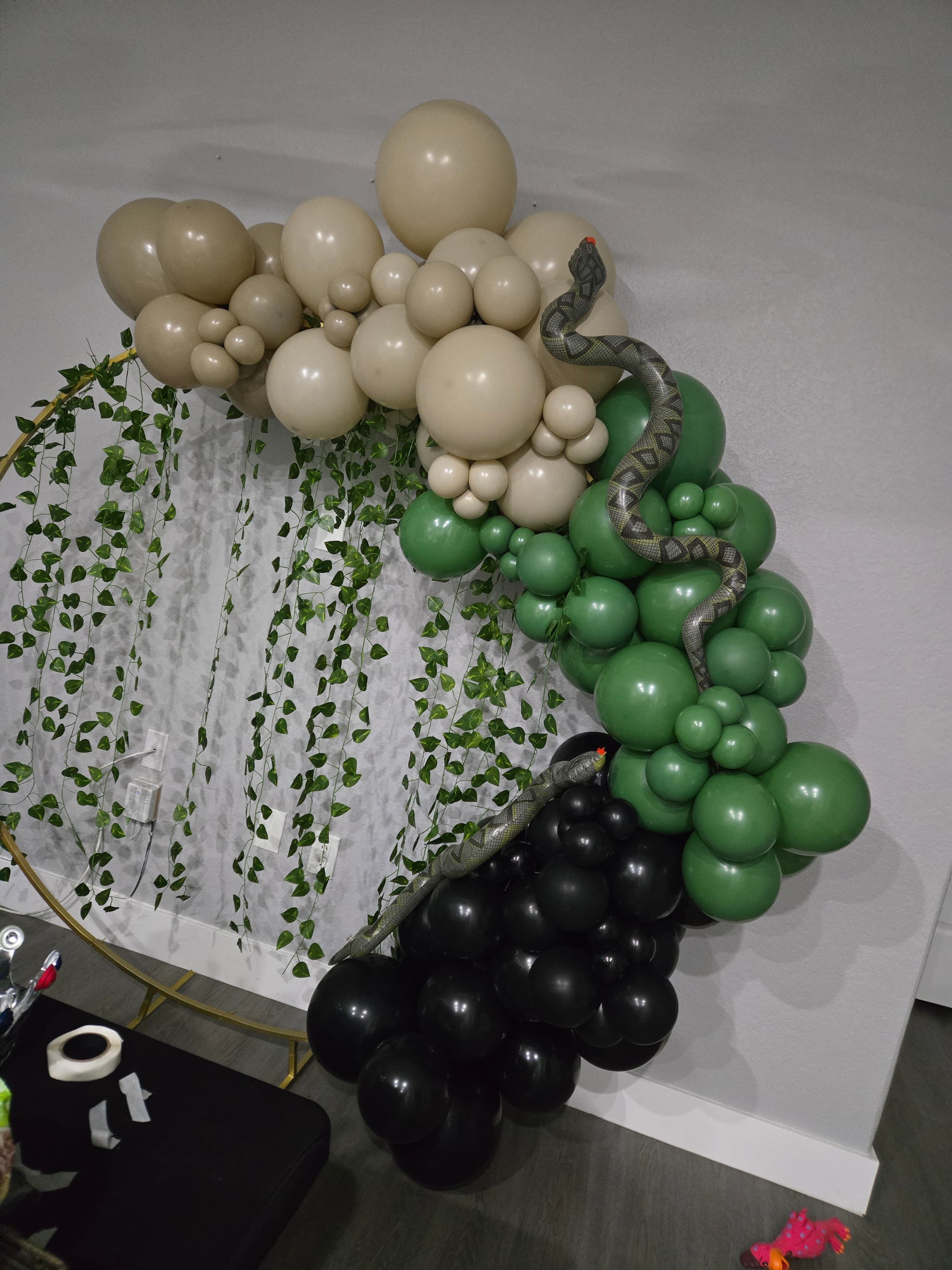 Tan, green, and black balloon garland decorated with toy snakes and hanging ivy vines.