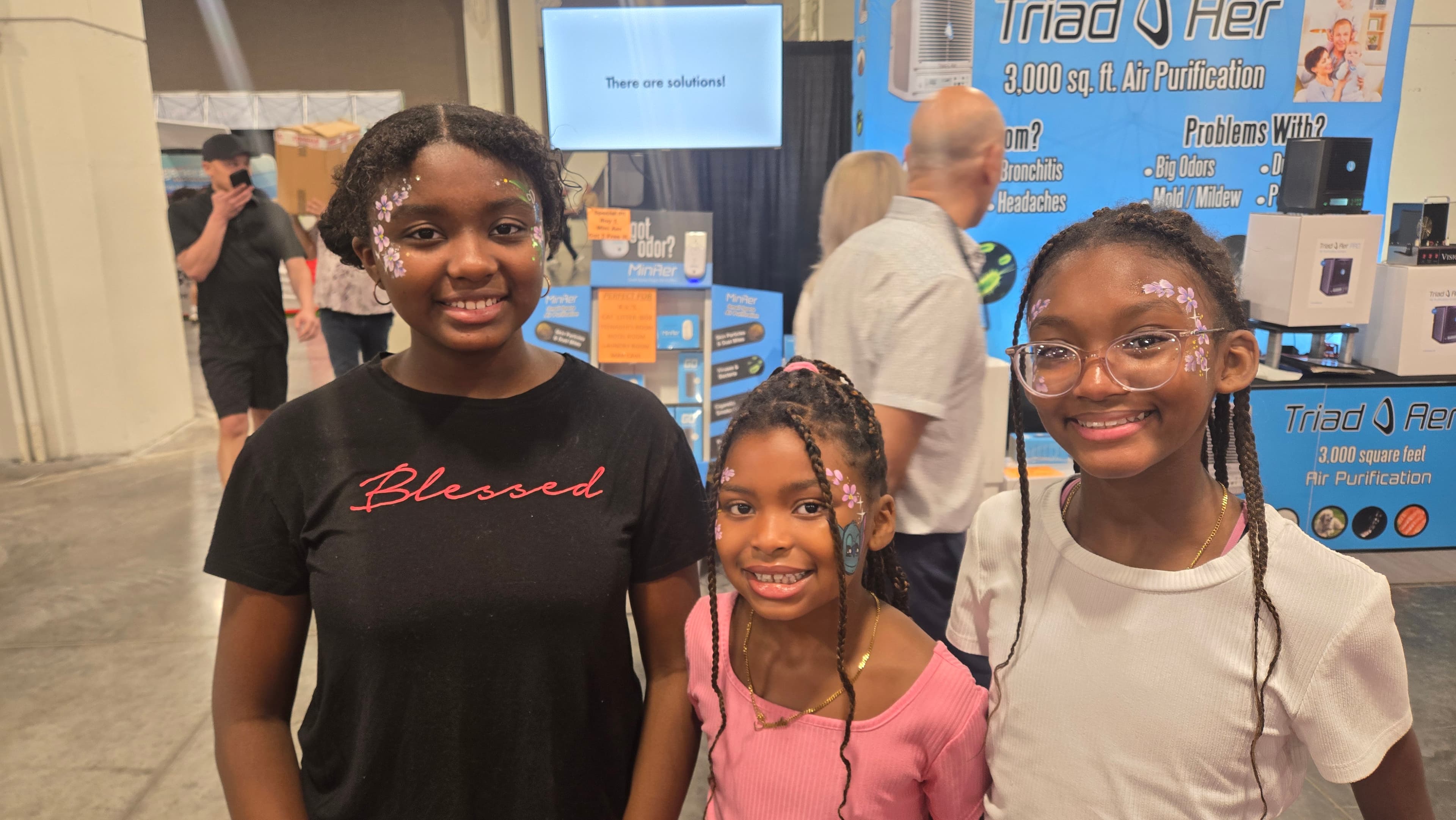 Three young Black girls with floral face paint smile at an indoor trade show.