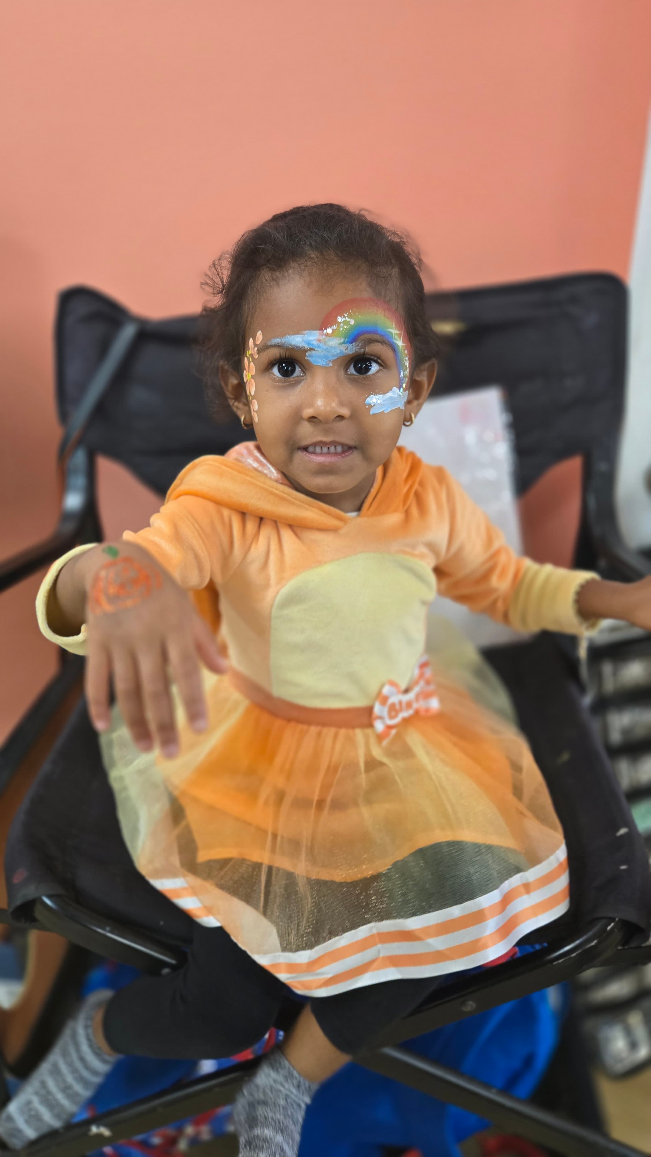 Smiling girl with rainbow face paint wearing an orange dress and reaching toward the camera.