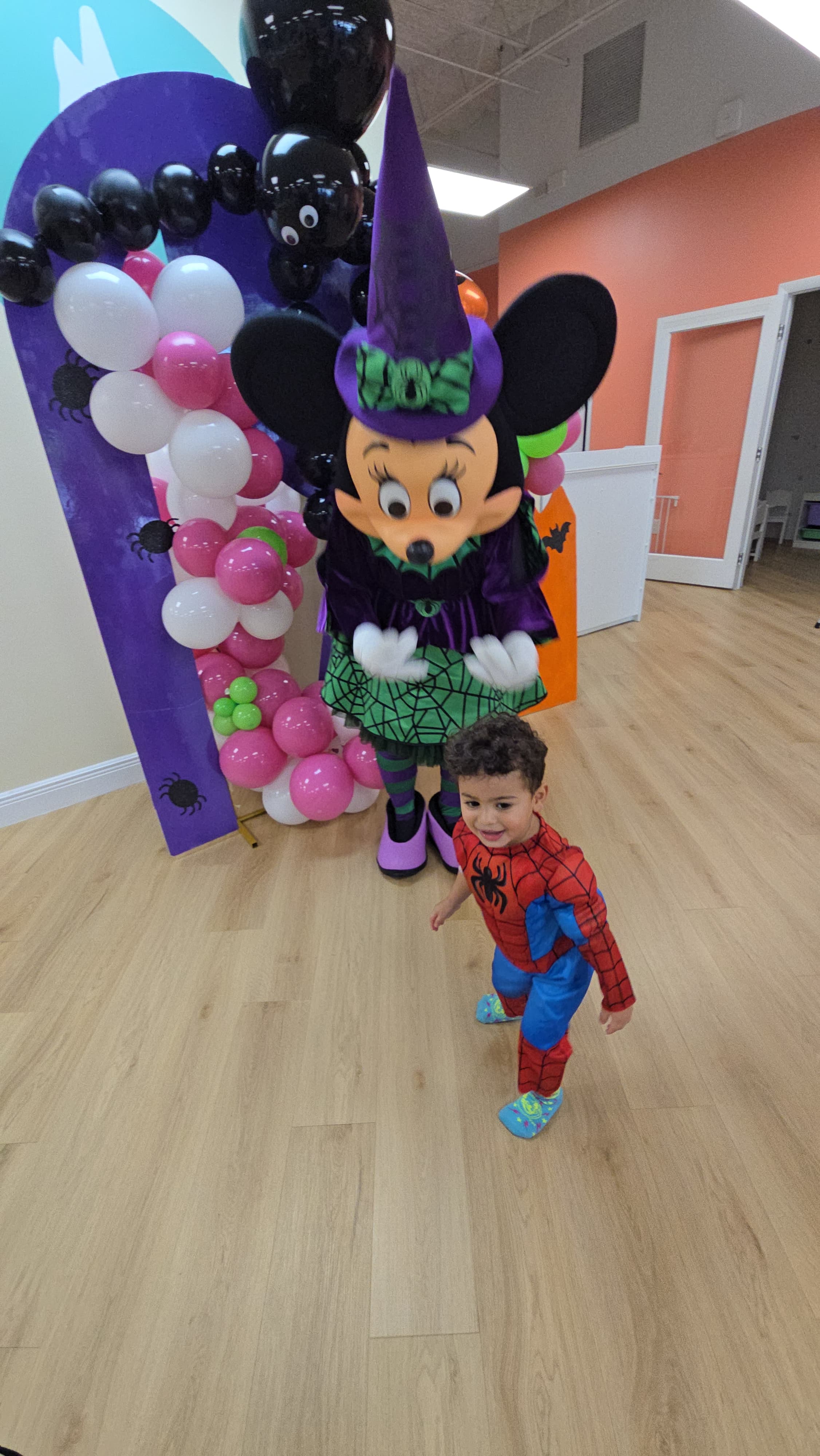 Young boy in Spider-Man costume smiles next to a Minnie Mouse witch mascot with balloons.