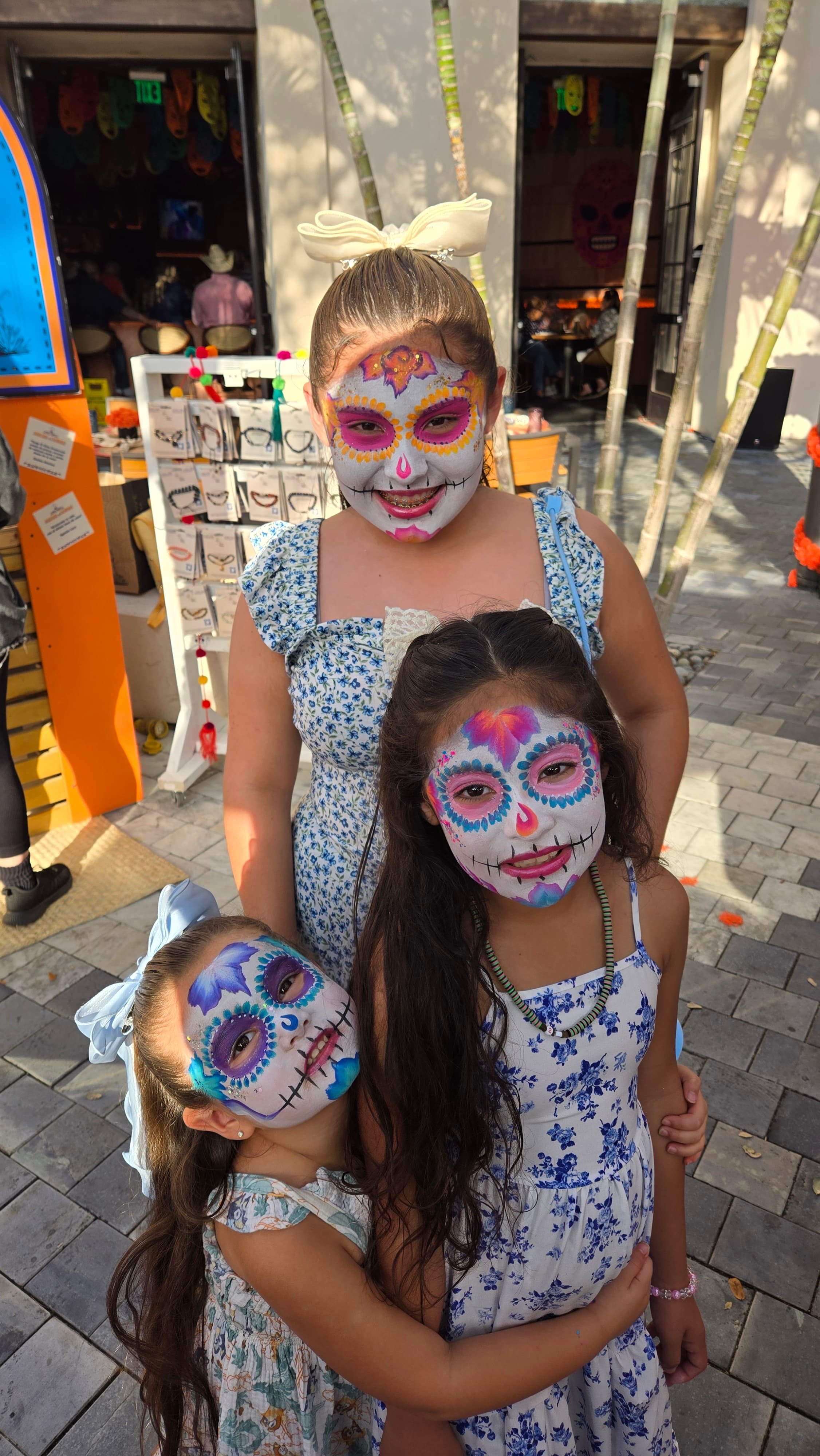 Three girls with vibrant sugar skull face paint and floral dresses pose together outside.