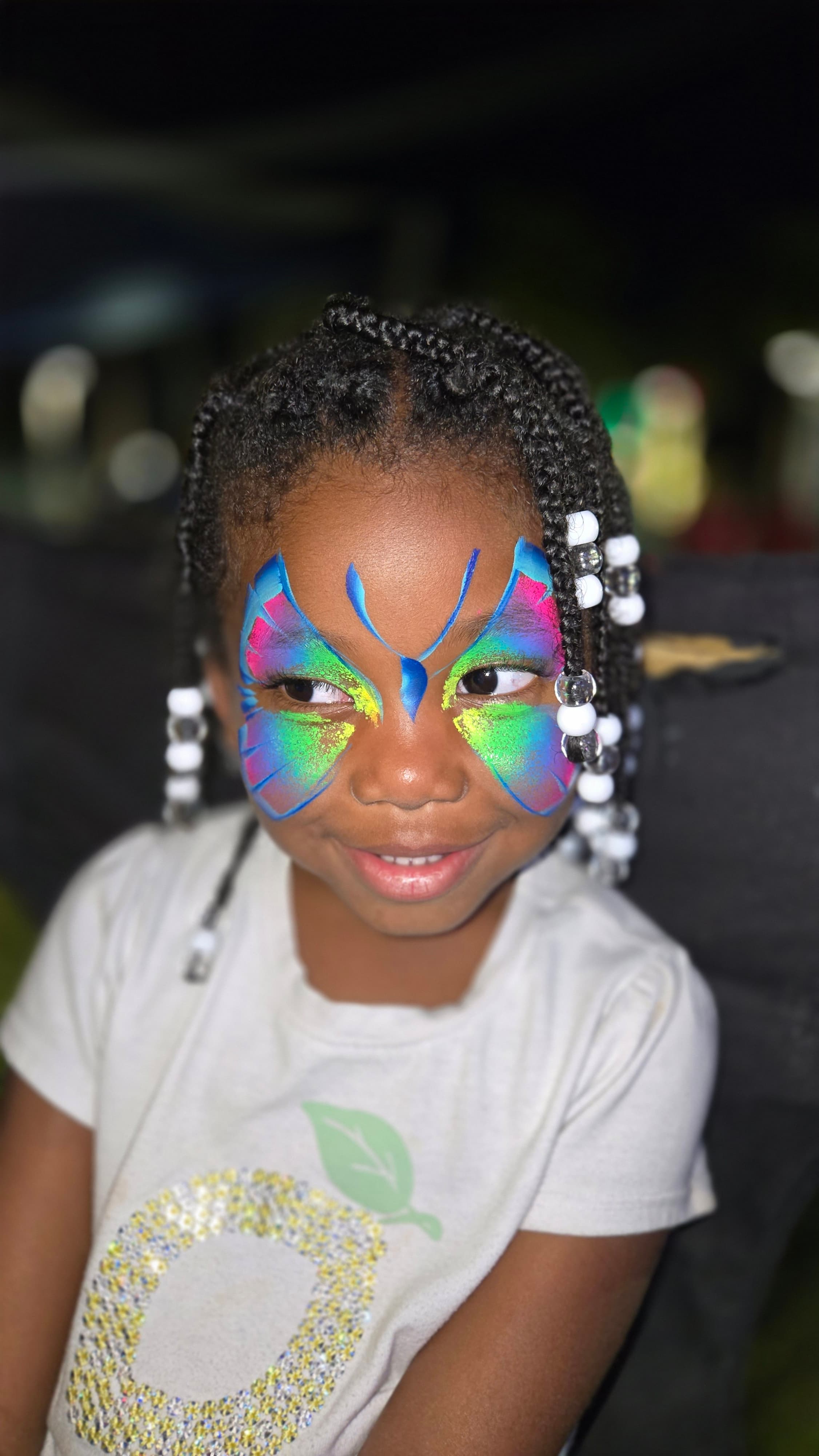 Smiling young girl with braided hair wearing vibrant blue, pink, and green butterfly face paint.