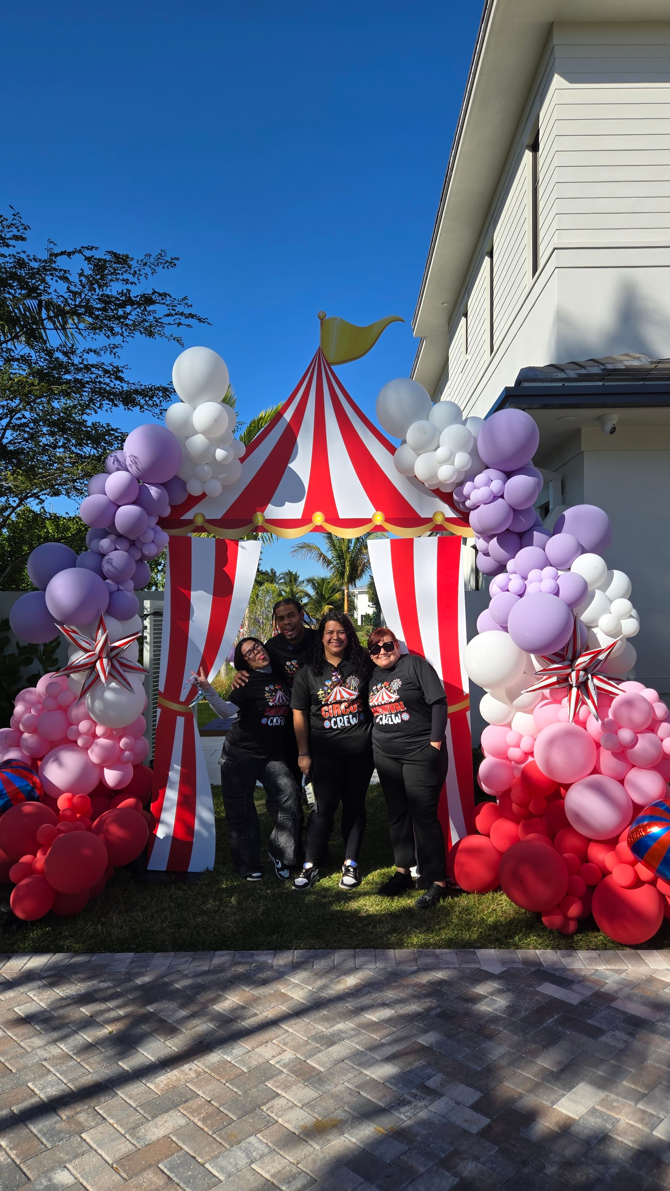 Four people pose under a red and white circus tent archway framed by colorful balloons.