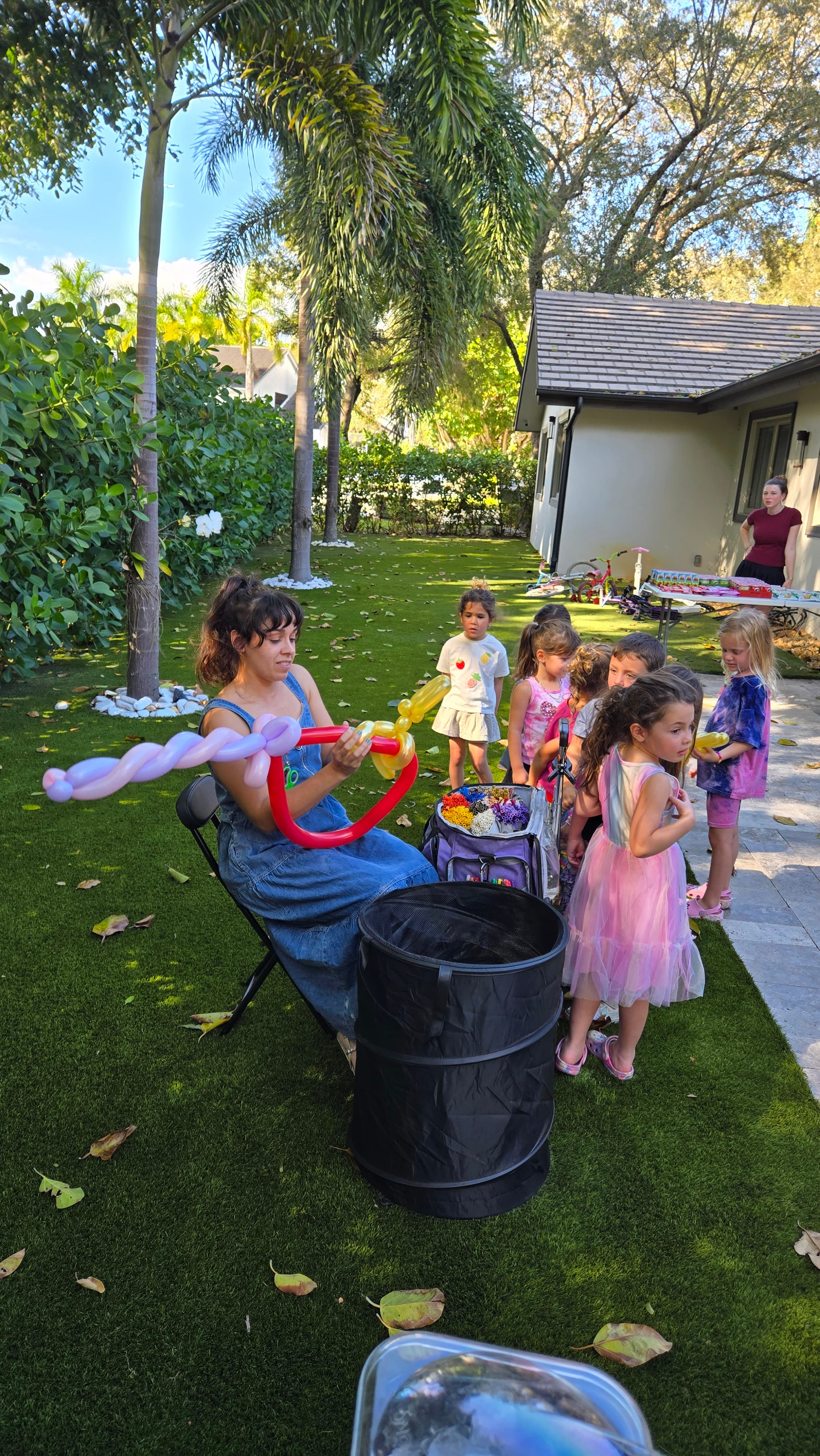 Woman in denim jumpsuit makes balloon animals for children on a sunny backyard lawn.