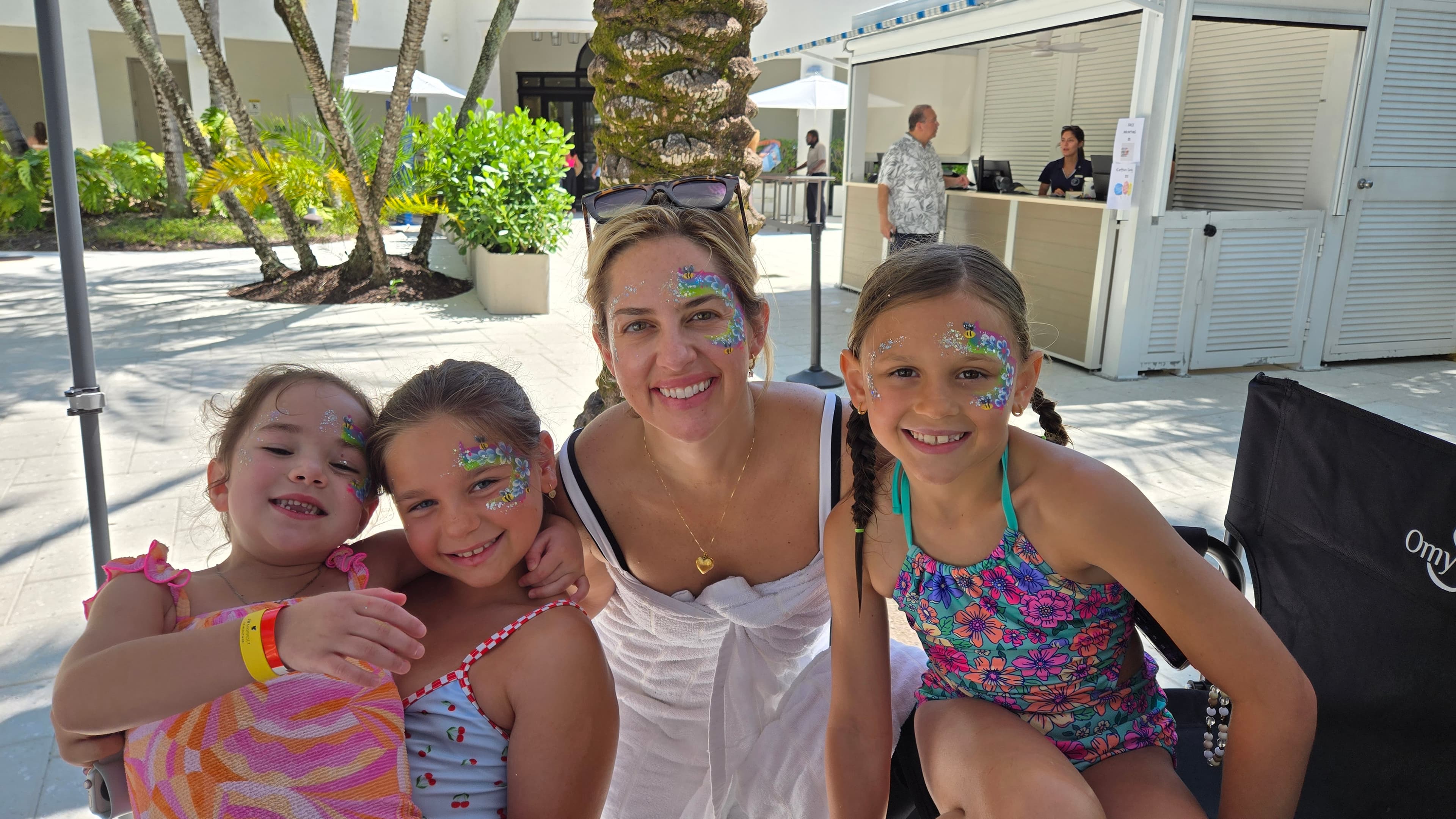 Smiling woman and three girls with colorful face paint pose together at a sunny resort.