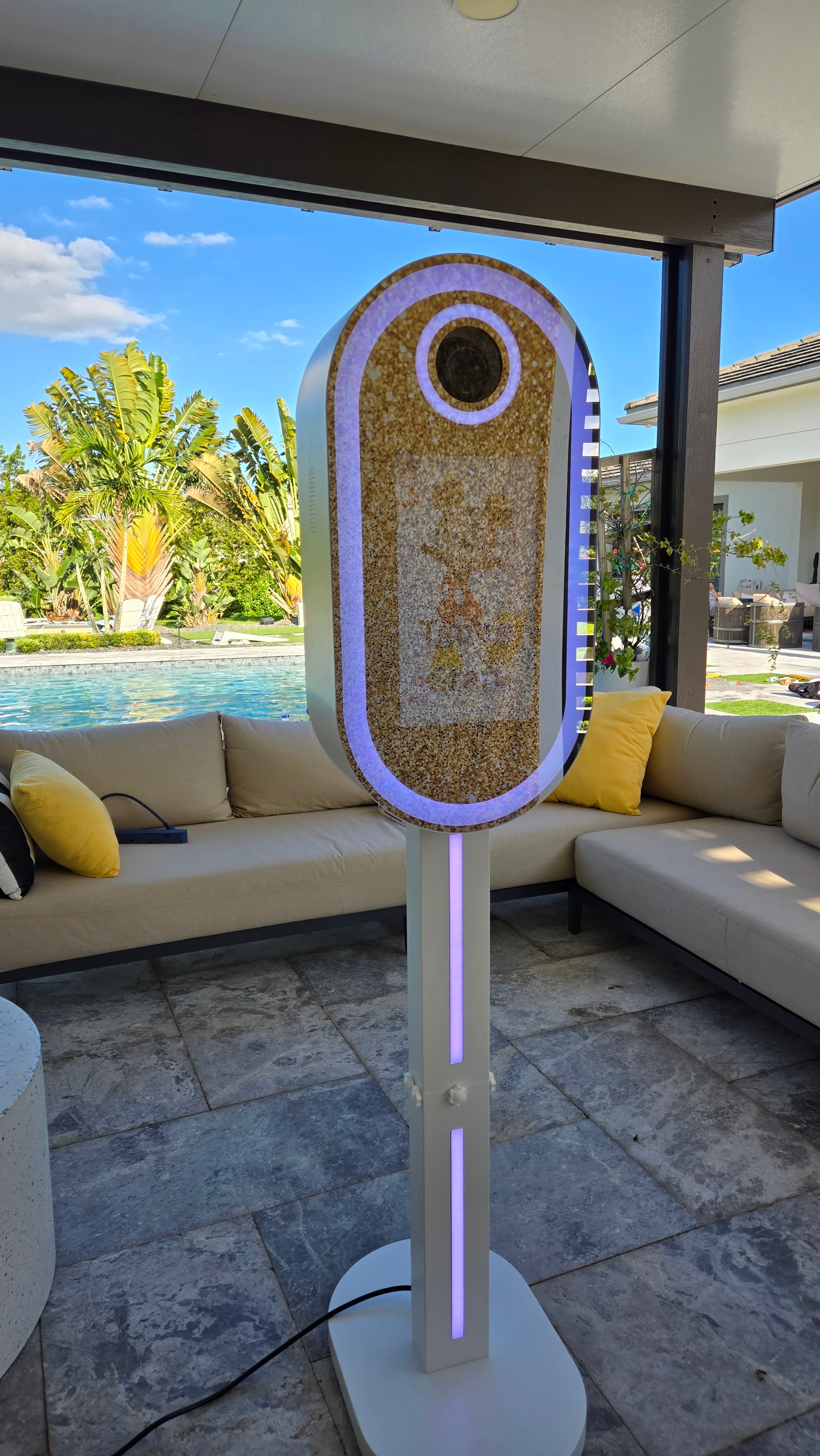 Oval gold glitter photo booth with purple LED ring light on a sunny patio.