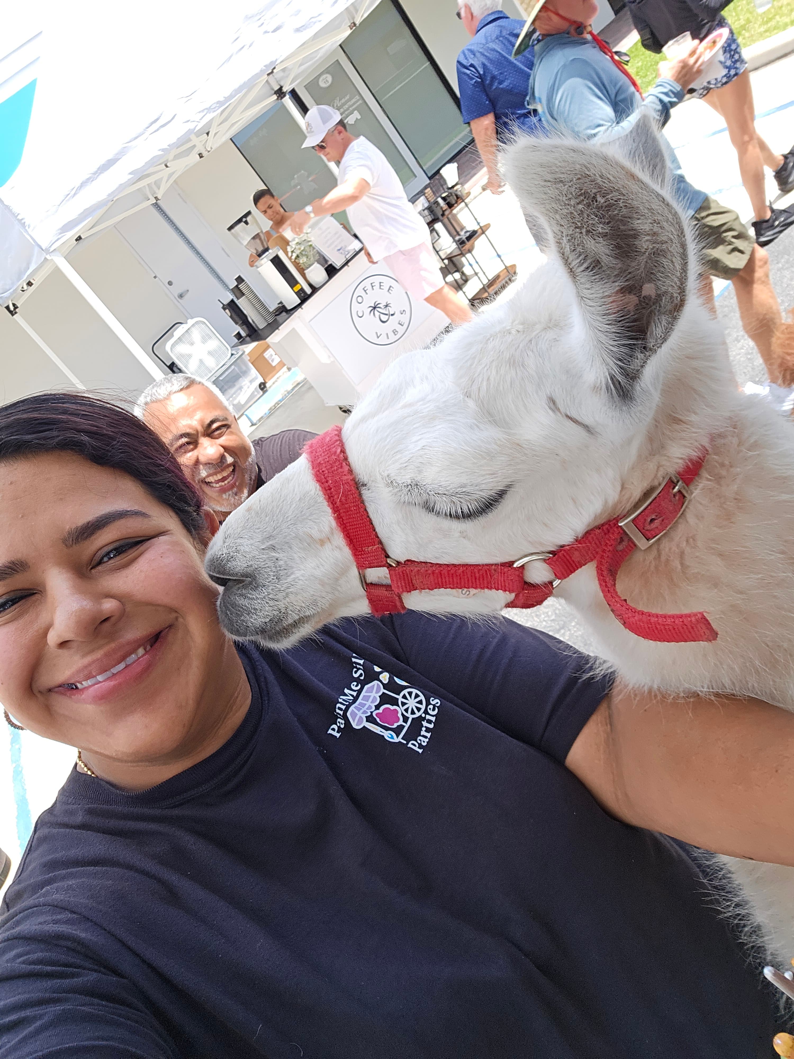 Smiling woman takes a selfie while a white llama in a red harness nuzzles her cheek.