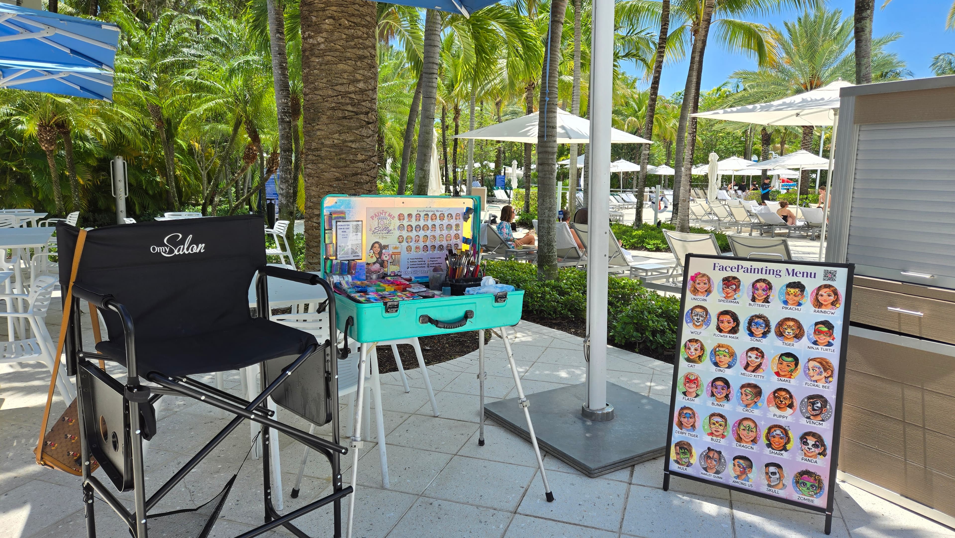 Face painting station with a teal kit and menu board at a sunny tropical resort.