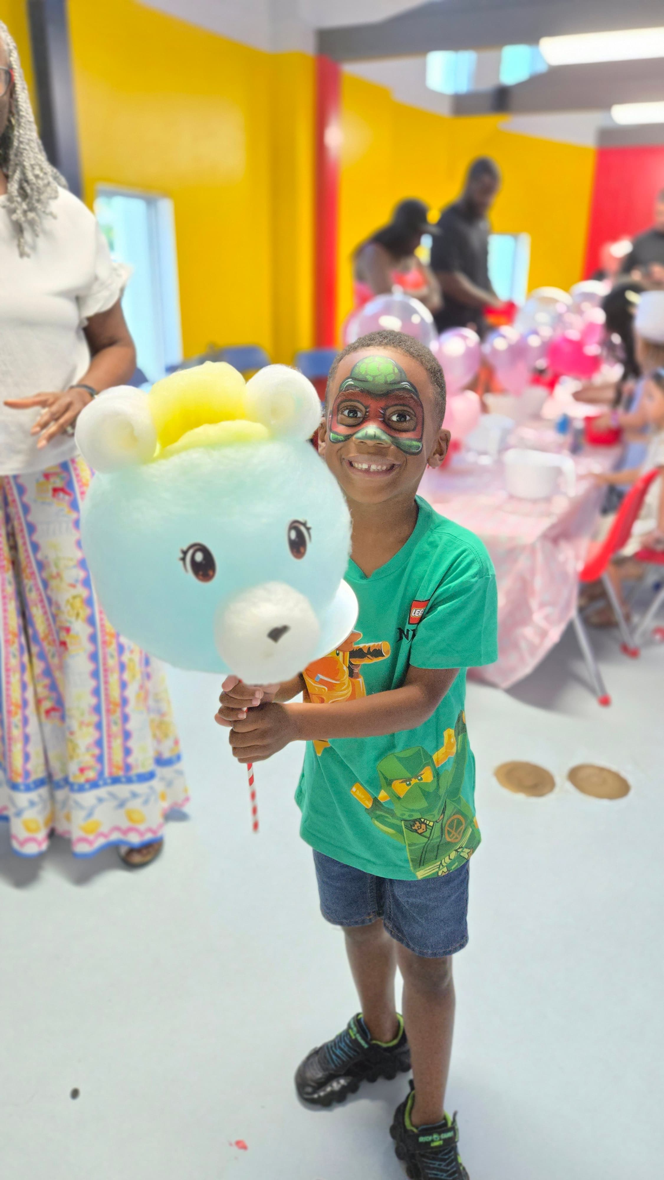 Smiling boy with turtle face paint holds a large, blue bear-shaped cotton candy treat.