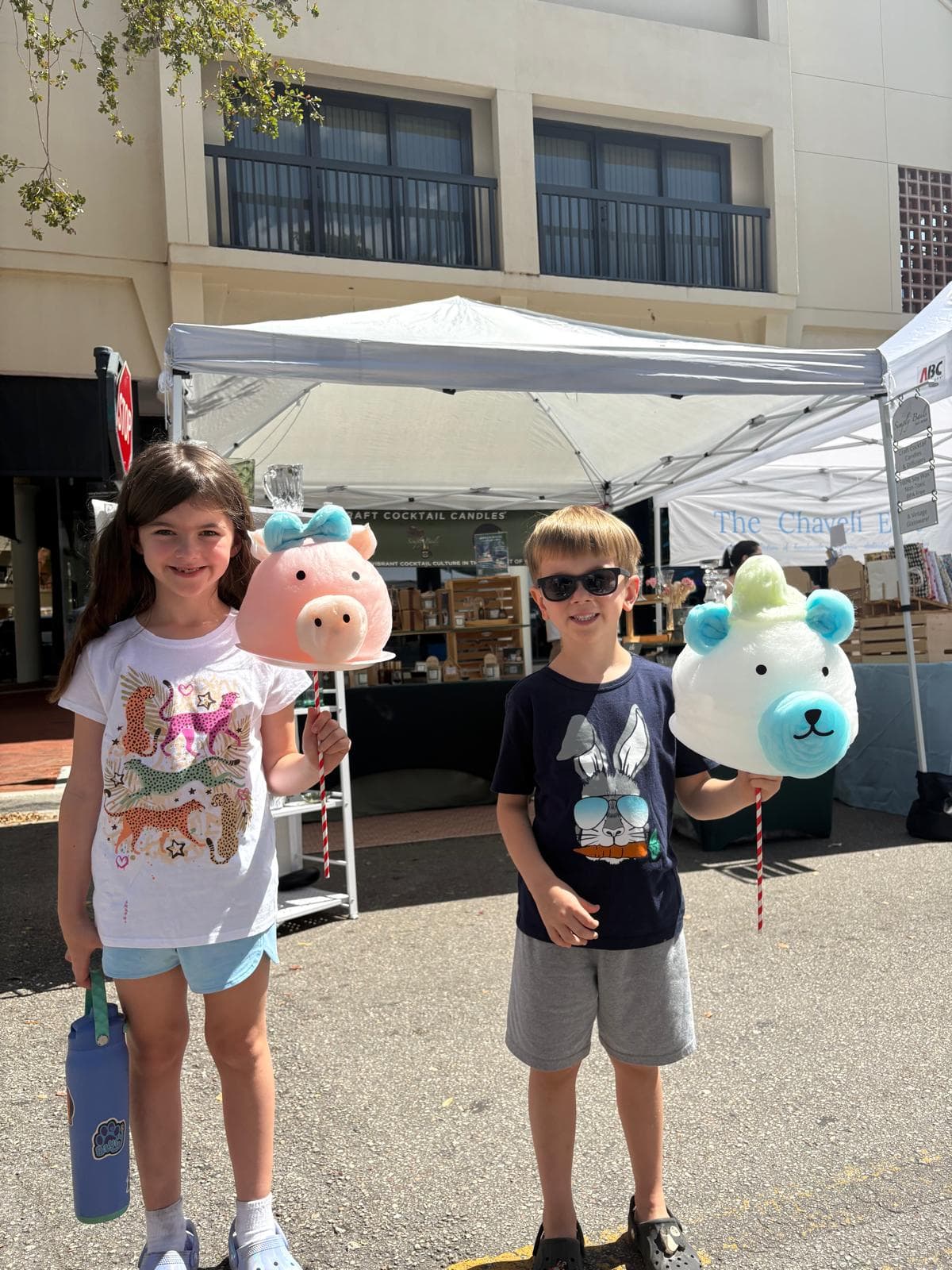 Two children holding pig and bear-shaped cotton candy at an outdoor market on a sunny day.