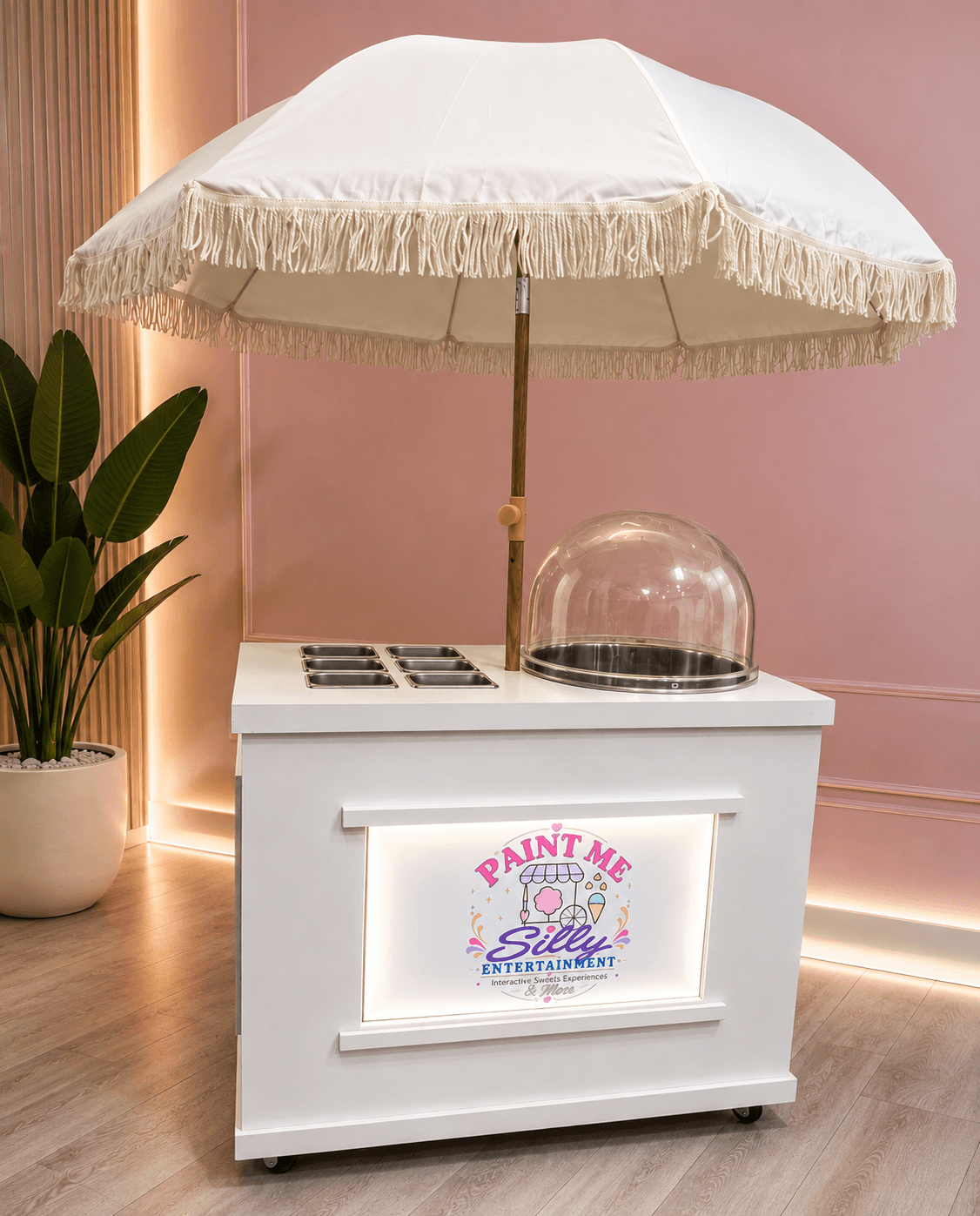 White mobile dessert cart with a fringed umbrella and cotton candy machine against pink walls.